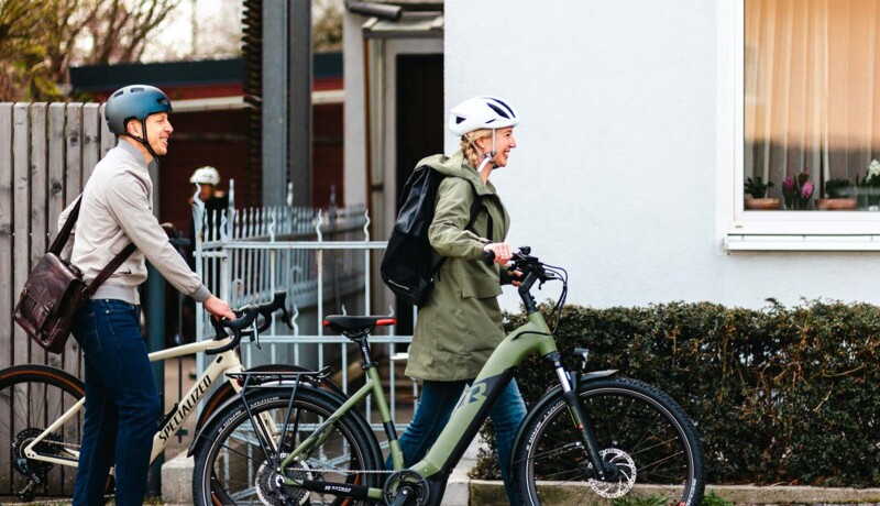 A man and a woman, both wearing bicycle helmets, are pushing their bicycles along a sidewalk. The woman is carrying a backpack, and both appear to be on their way somewhere.
